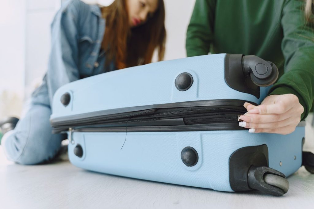 From below of crop concentrated young woman in denim outfit closing suitcase sitting on floor with anonymous friend while preparing for trip