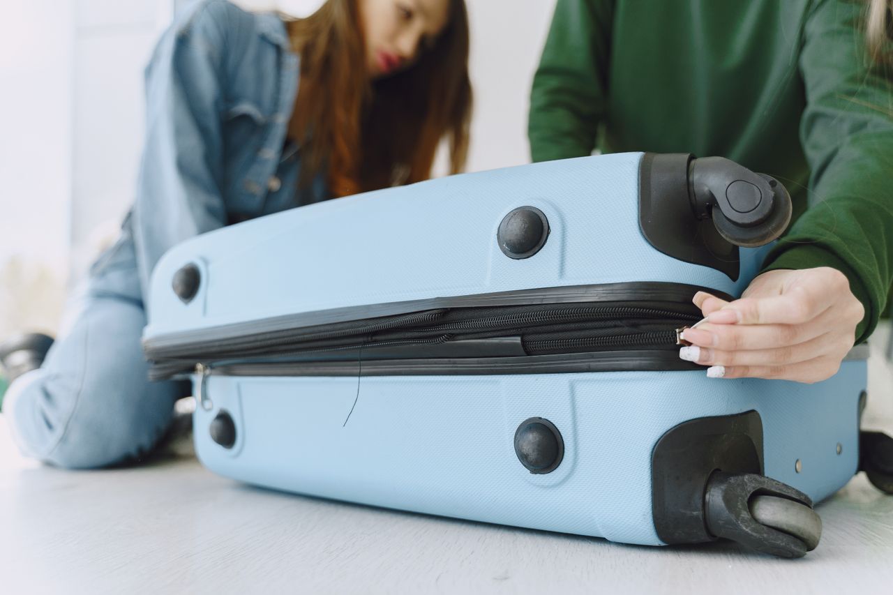 From below of crop concentrated young woman in denim outfit closing suitcase sitting on floor with anonymous friend while preparing for trip